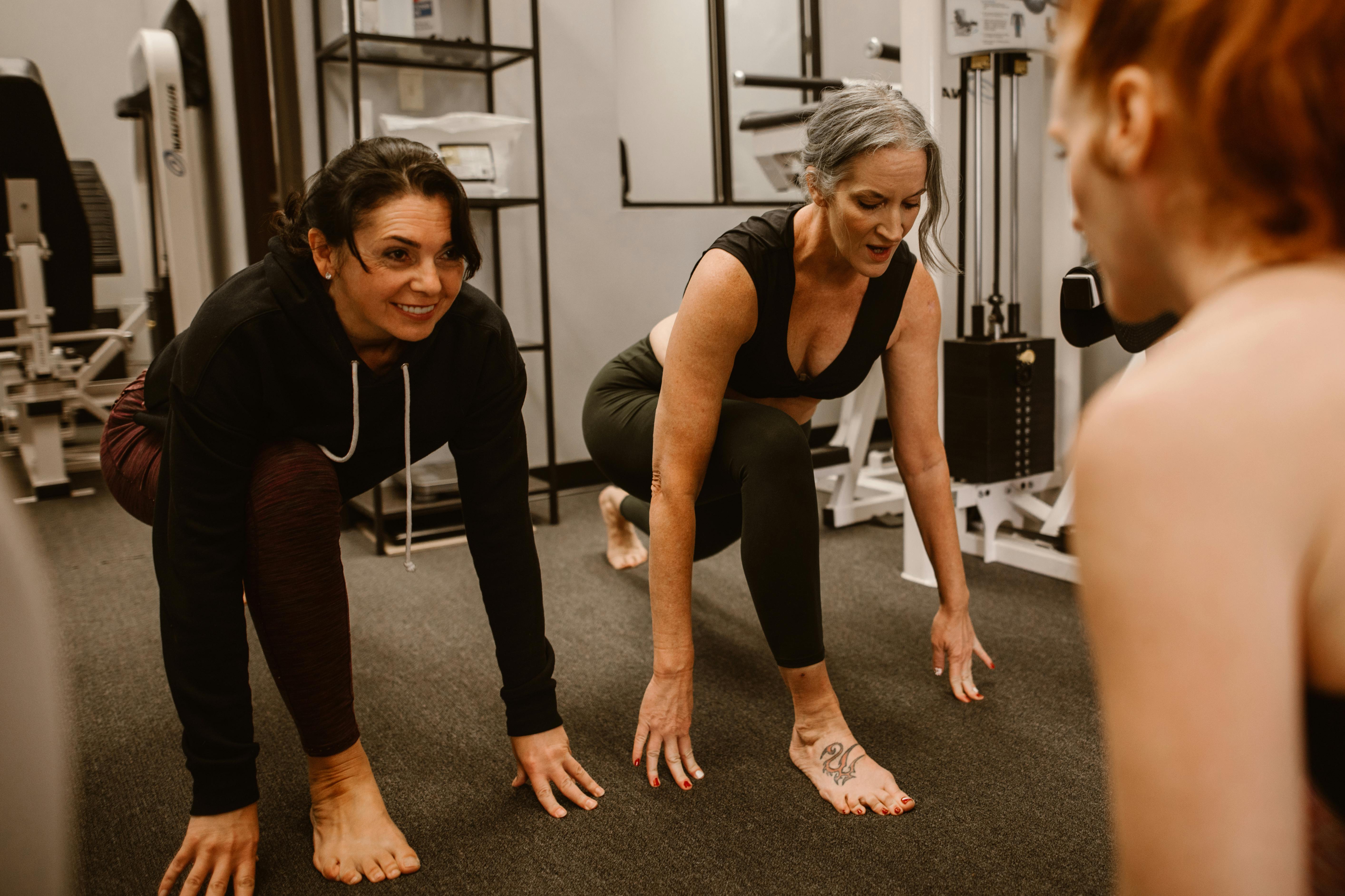 Women stretching in a gym during a fitness class, barefoot in lunge position, guided by a trainer.